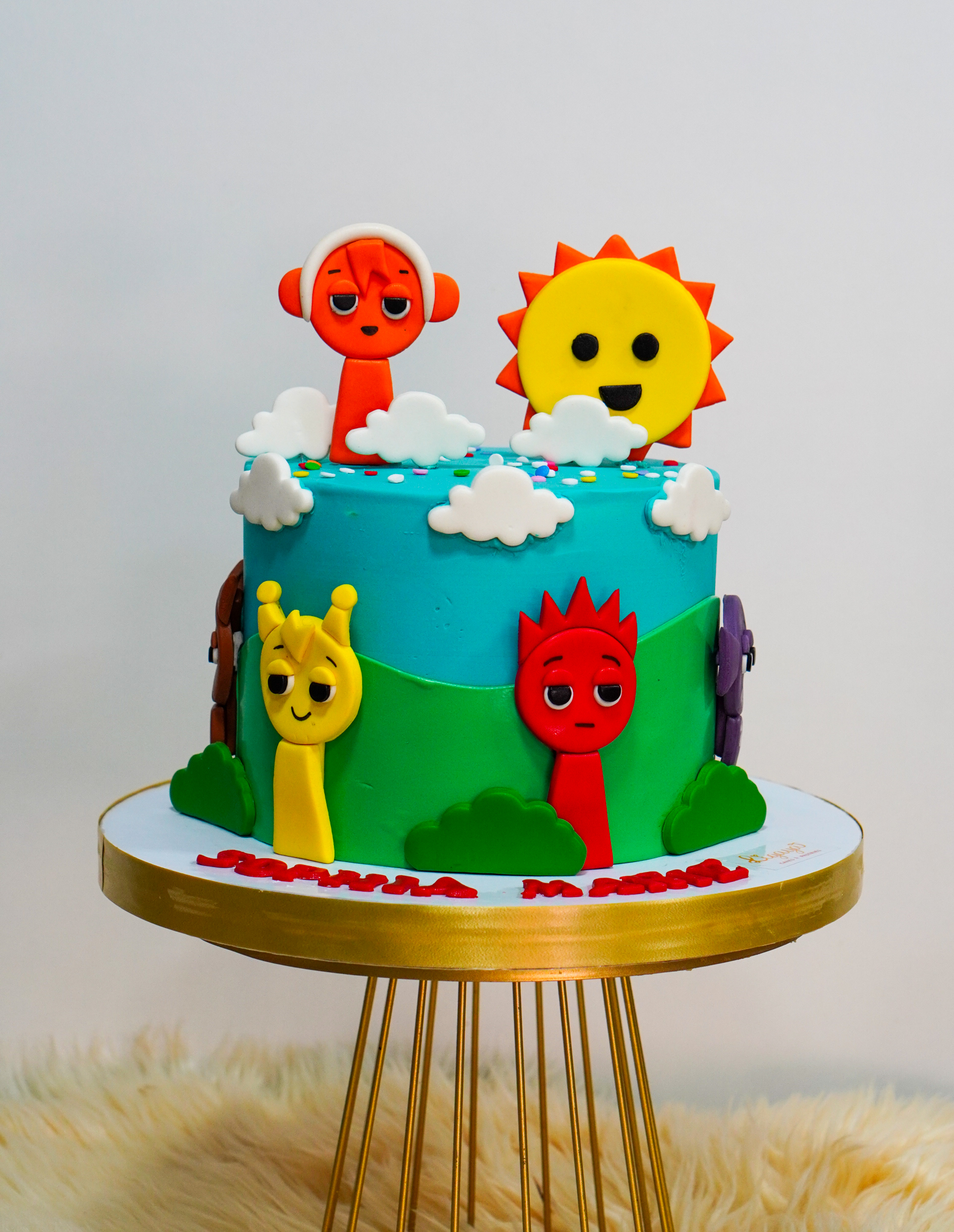 Colorful fondant birthday cake with cartoon sun and animal characters on a grassy landscape background