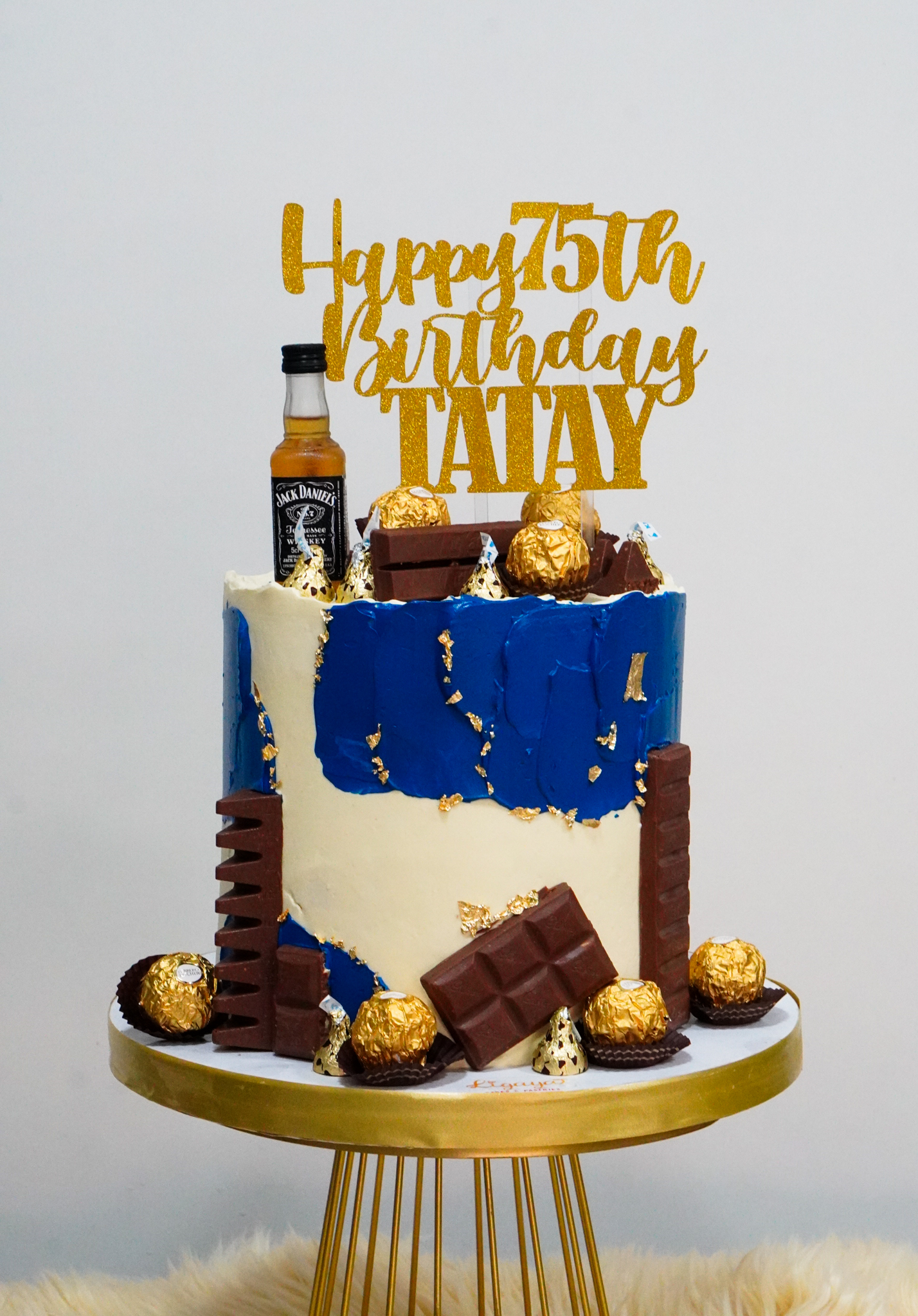 Blue and white buttercream birthday cake with gold flakes, topped with Jack Daniel's miniature bottle, Ferrero Rocher chocolates, and chocolate bars