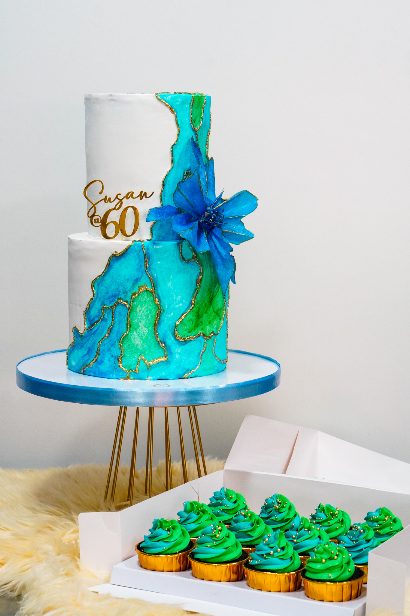 Elegant white and blue geode-inspired two-tier birthday cake with a large blue flower, paired with matching green and blue cupcakes