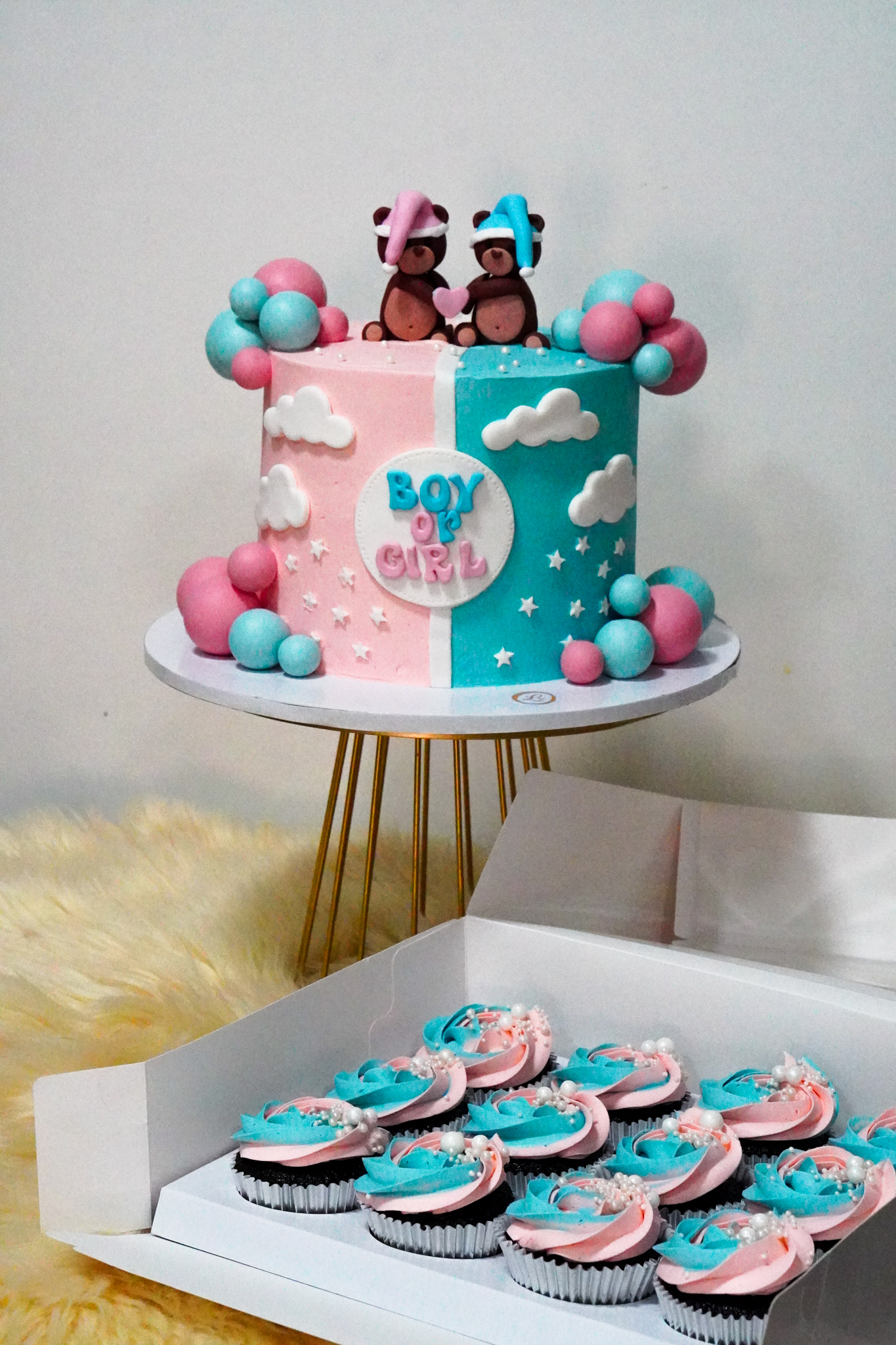 Gender reveal cake in pastel pink and blue with fondant clouds, stars, and balloon clusters, topped with two teddy bears wearing pink and blue hats, and matching pink and blue frosted cupcakes with pearl sprinkles in a box