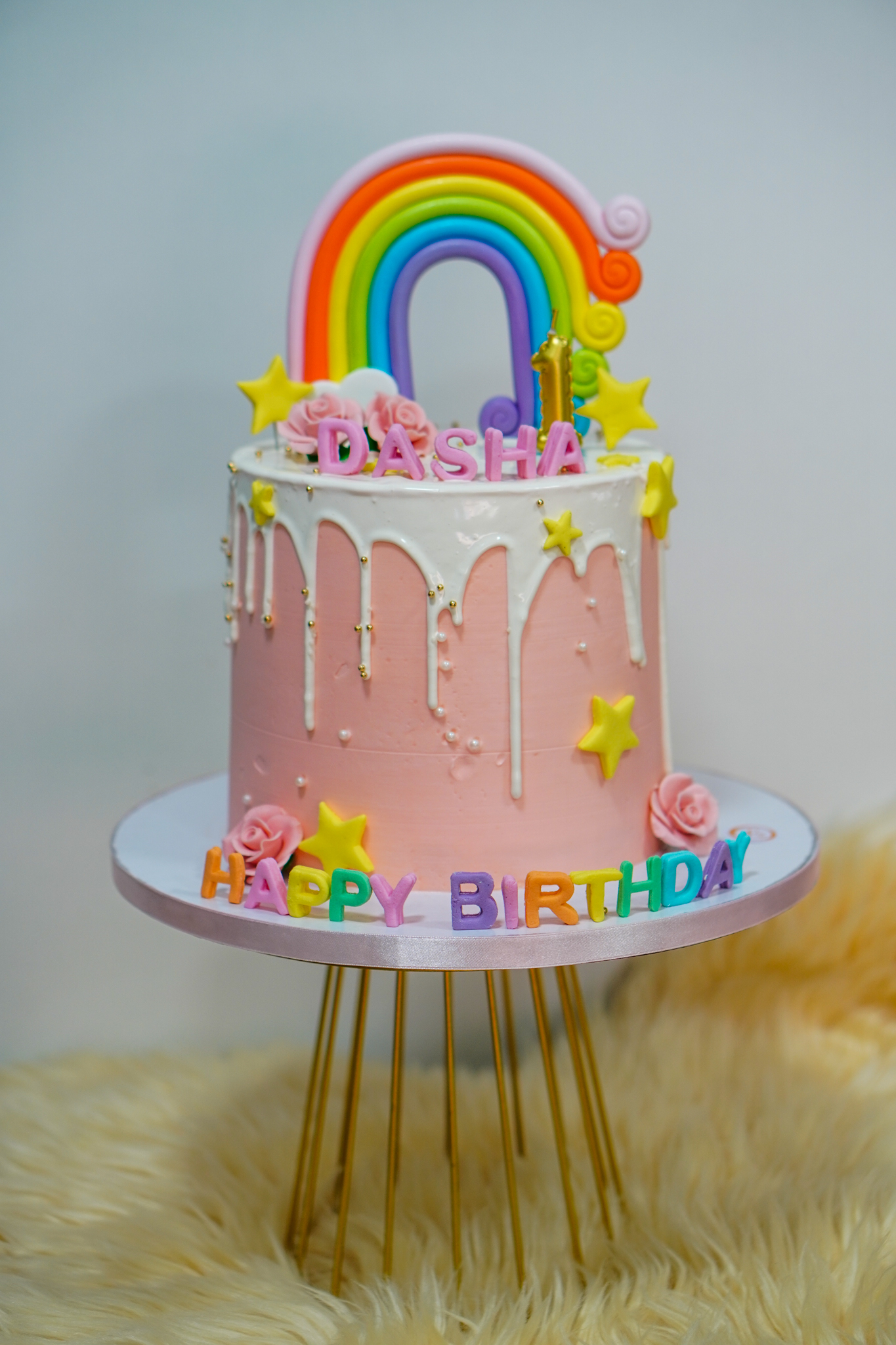 Pink buttercream birthday cake with white drip design, decorated with a fondant rainbow topper, stars, roses, and colorful 'Happy Birthday' letters, featuring the name Dasha