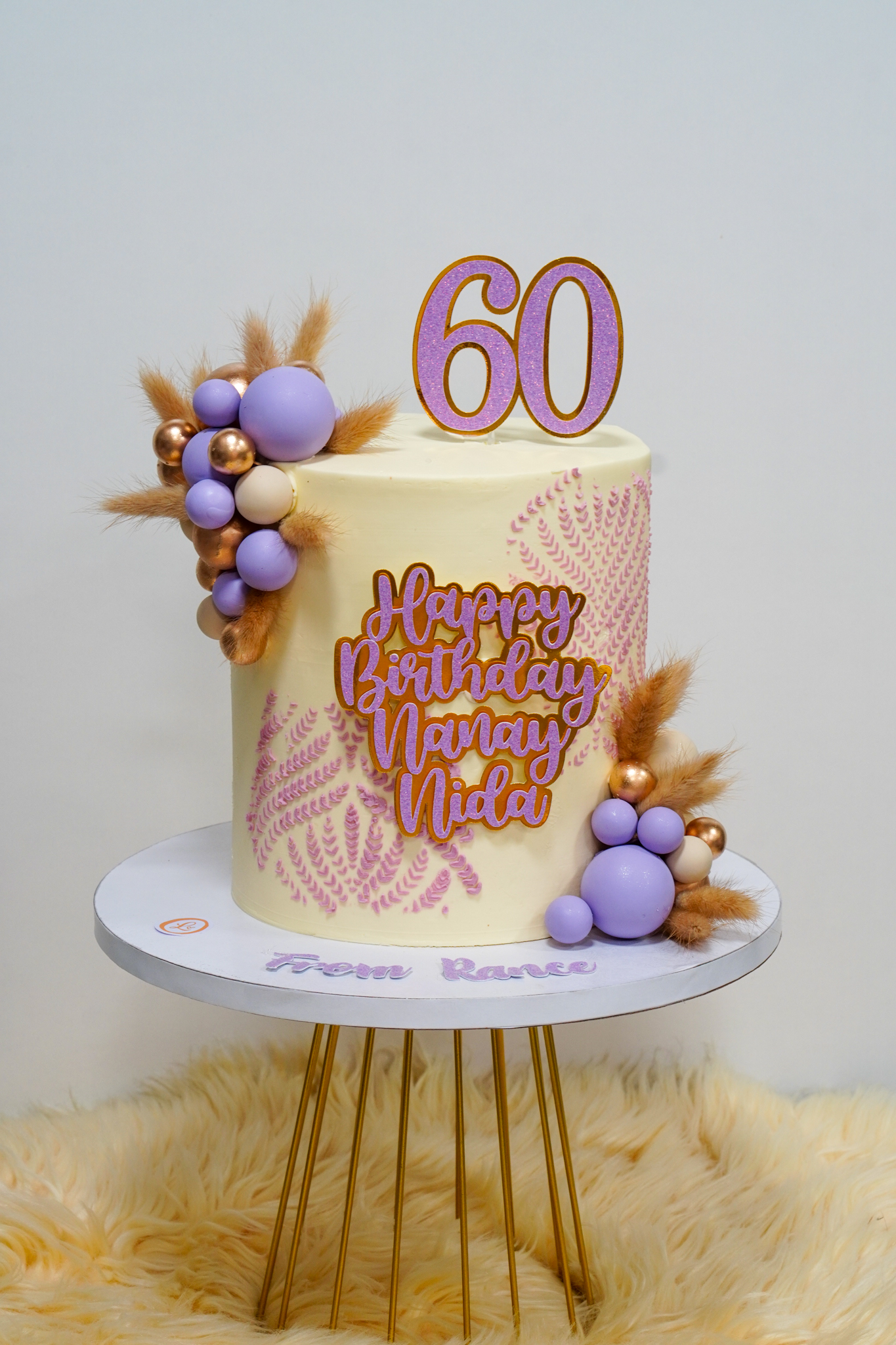 Tall cream-colored birthday cake with pink fern patterns, purple and gold decorative balls, brown dried floral accents, and a large '60' cake topper