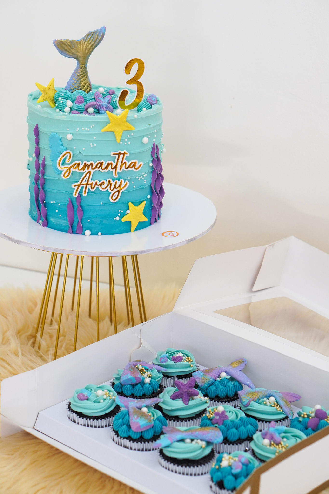 Mermaid themed blue buttercream birthday cake with gold mermaid tail topper, yellow starfish, number 3 topper, and matching ocean-themed cupcakes decorated with tails and seashells