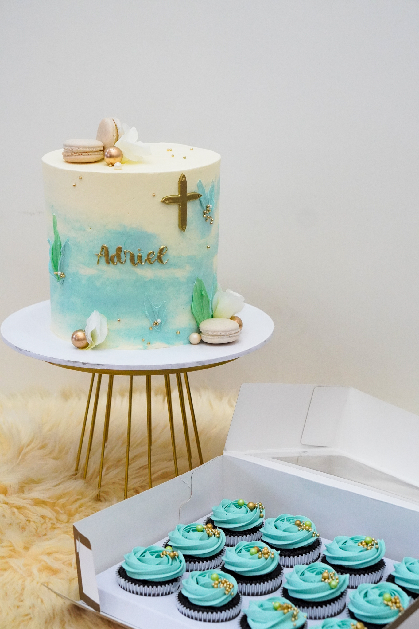 Blue and cream buttercream cake with gold cross, green leaf decorations, macarons, gold pearls, and the name 'Adriel', accompanied by matching blue frosted cupcakes