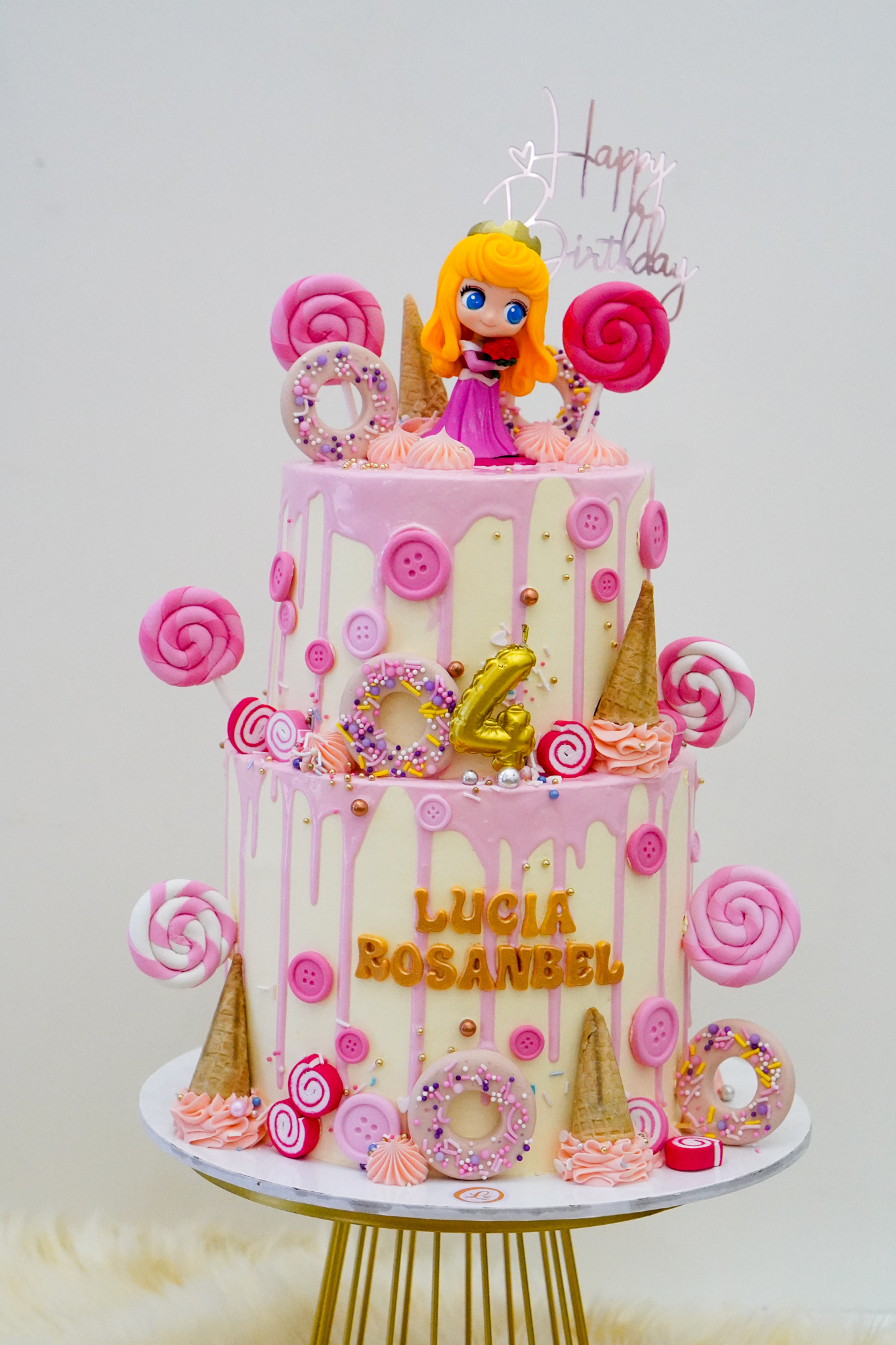 Two-tier pink and cream buttercream birthday cake with pink drip design, topped with a doll figurine, large lollipops, donuts, ice cream cones, and the text 'Lucia Rosanbel' with a gold number 4 topper