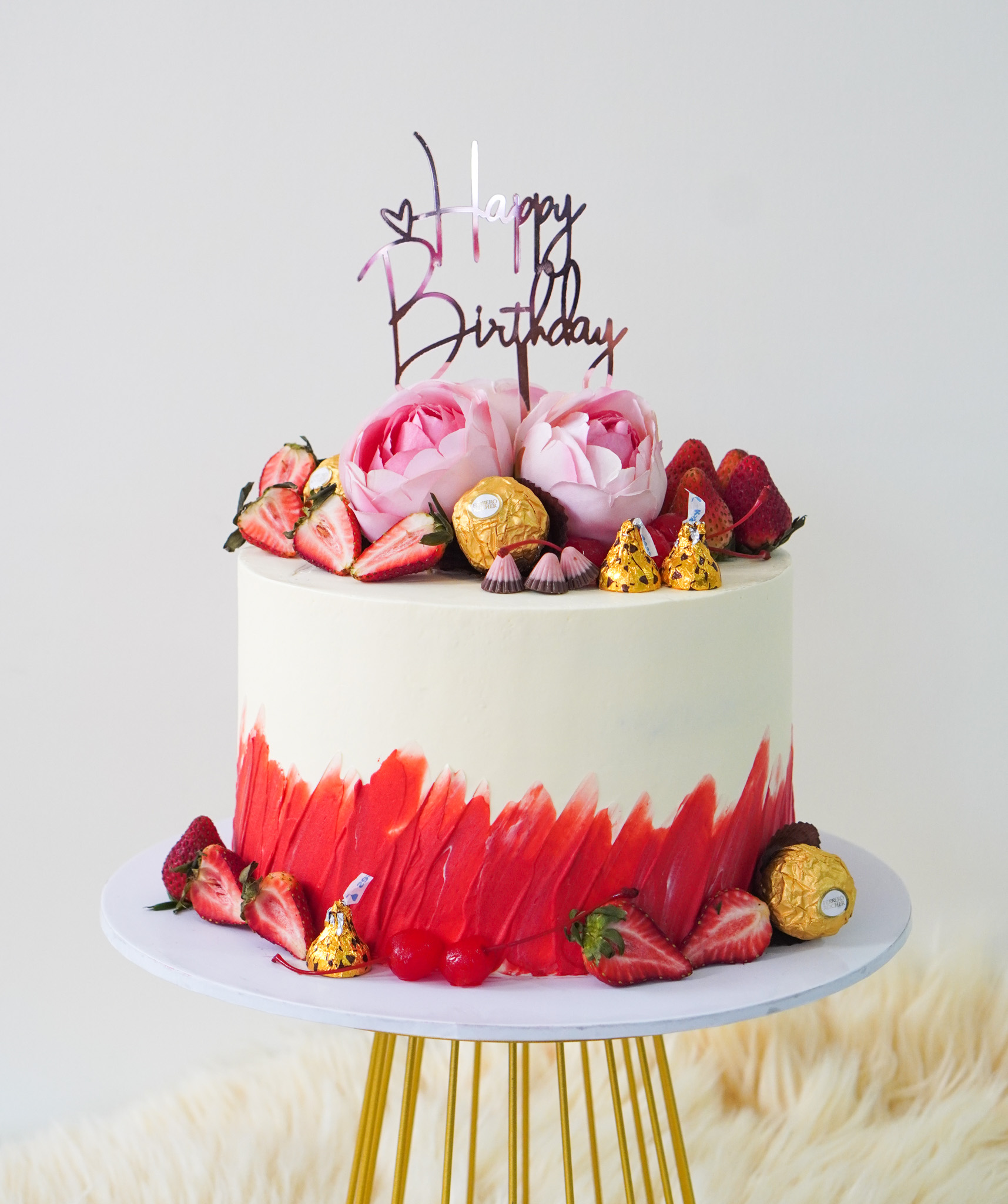 Cream buttercream birthday cake with red brushstroke design, topped with pink flowers, strawberries, cherries, Ferrero Rocher chocolates, Hershey's Kisses, and a 'Happy Birthday' topper