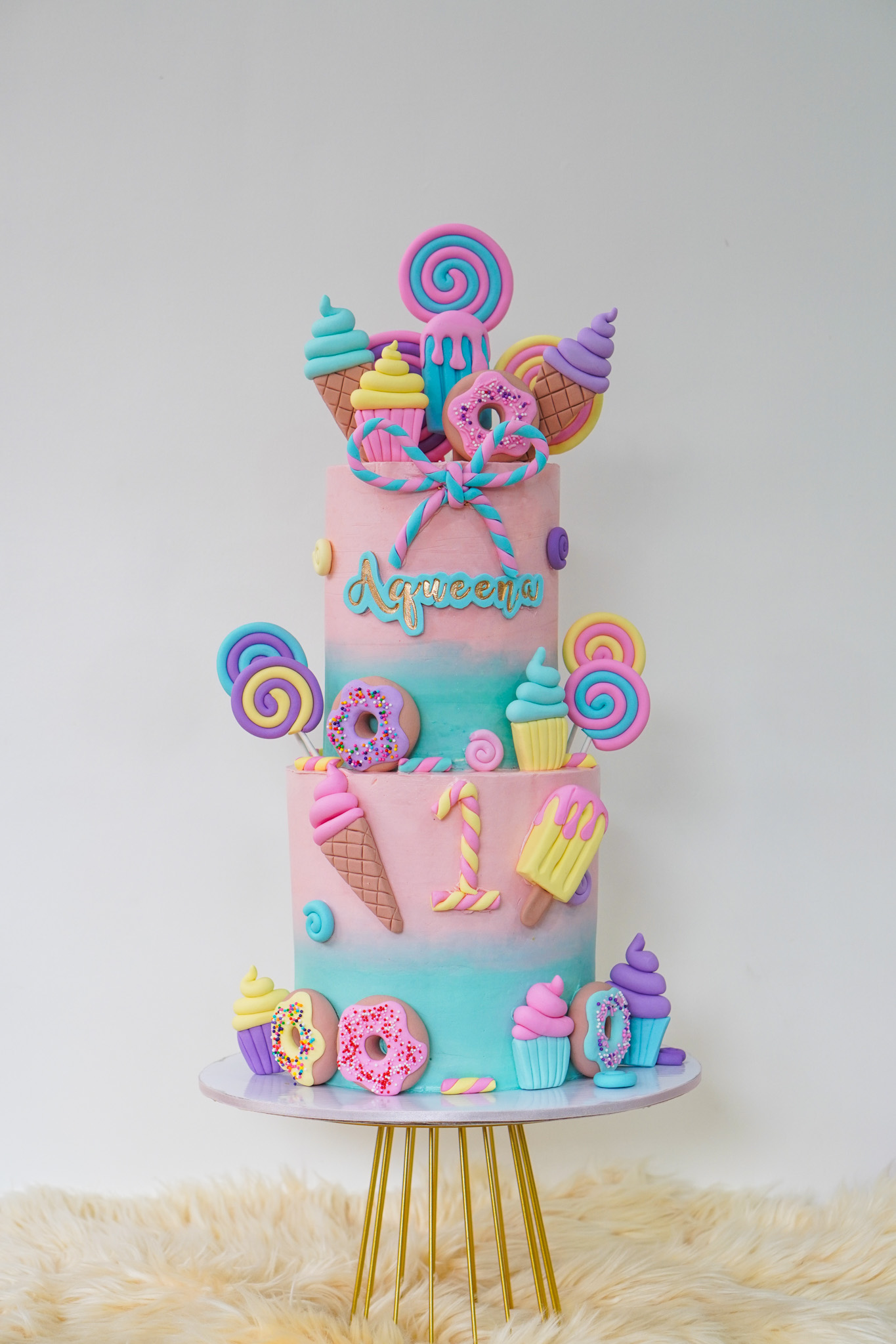 Two-tier pastel pink and blue ombré fondant cake decorated with ice cream cones, lollipops, donuts, cupcakes, popsicles, and the name 'Aqueena' with a number 1 topper